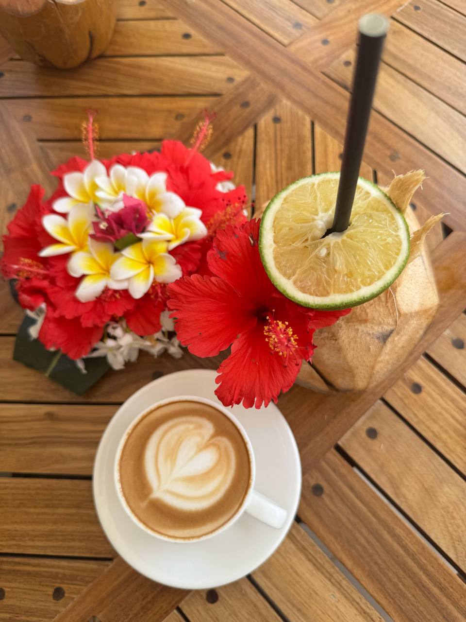 A cup of cappuccino with heart latte art, tropical flowers and a fresh coconut drink on a wooden table